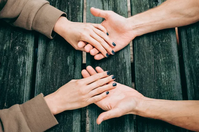 Vue de haut en bas de deux paires de mains se rencontrant sur une table en bois sombre et rustique. Une personne a du vernis à ongles bleu foncé/gris. Le geste est solidaire et intime, montrant un lien humain profond.