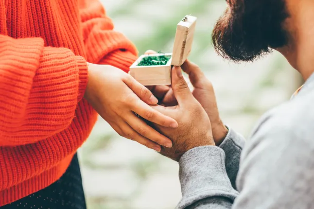 Gros plan des mains d'un homme présentant une petite boîte cadeau en bois à une femme vêtue d'un pull orange vif.