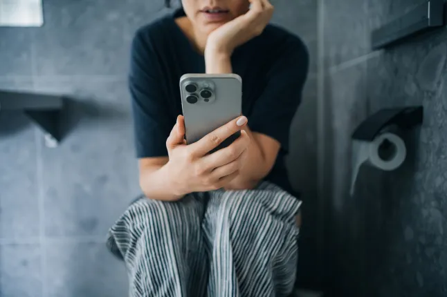 Plan recadré d'une jeune femme asiatique utilisant un smartphone alors qu'elle était assise sur les toilettes dans la salle de bain. Routine quotidienne. Style de vie et technologie