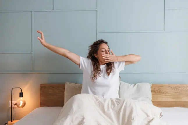 Une femme d'âge moyen aux cheveux bouclés bâille et s'étire en étant assise dans son lit. Elle porte un t-shirt blanc et est entourée d'une literie neutre dans une chambre moderne doucement éclairée.