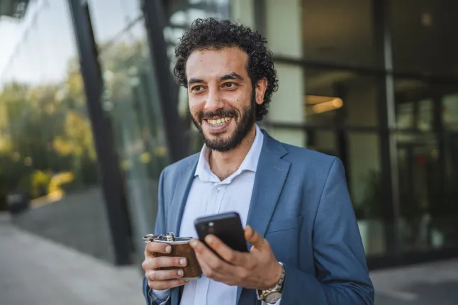 Jeune manager en costume élégant, tenant une flasque et un téléphone intelligent, souriant avec confiance tout en détournant le regard des immeubles de bureaux modernes dans un environnement urbain pendant la journée