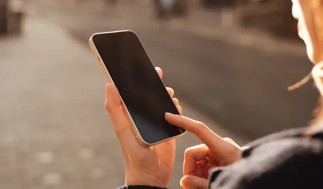 Main féminine défilant sur un téléphone intelligent, avec une rue de ville légèrement floue derrière elle dans la chaude lumière du soleil. Connexion et présence dans la vie urbaine moderne. Concept de contact humain, de technologie et de routine quotidienne.