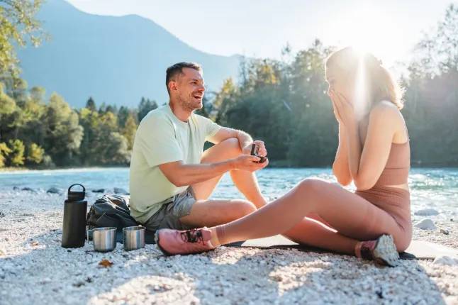 Un homme joyeux sur un genou propose à sa petite amie surprise par une journée ensoleillée au bord d'une rivière lors d'un voyage de camping. Excitation et amour dans la nature.