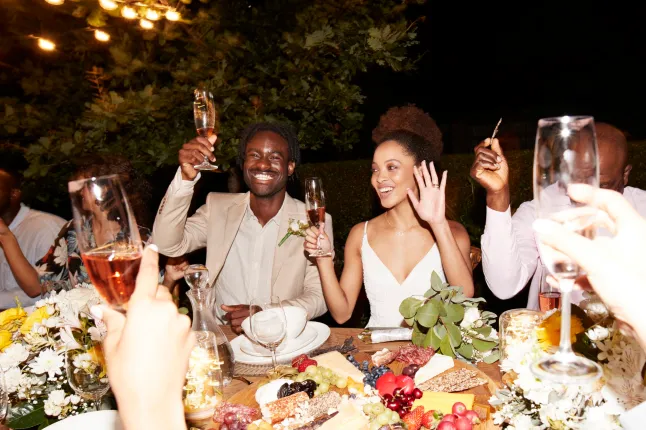 Joyeux jeune couple de jeunes mariés grillant des boissons avec les invités du mariage à une table à manger éclairée pendant la réception