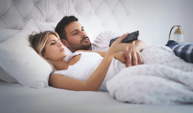 Vue latérale rapprochée d'un couple d'une vingtaine d'années profitant d'un matin de week-end paresseux en regardant la télévision au lit. Ils sont confortablement installés et surfent sur les chaînes. La femme tient la télécommande.