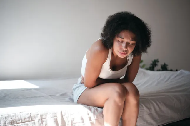 Symptômes du syndrome prémenstruel. Une jeune femme afro-américaine touchant le ventre souffre de maux d'estomac, assise sur son lit à la maison, de douleurs abdominales et d'estomac