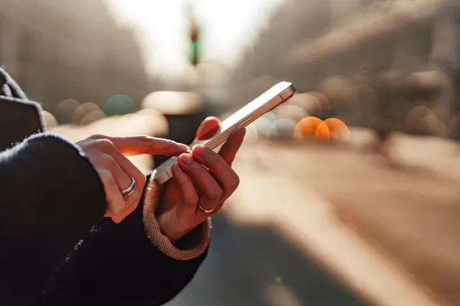 Main féminine défilant sur un téléphone intelligent, avec une rue de ville légèrement floue derrière elle dans la chaude lumière du soleil. Connexion et présence dans la vie urbaine moderne. Concept de contact humain, de technologie et de routine quotidienne.
