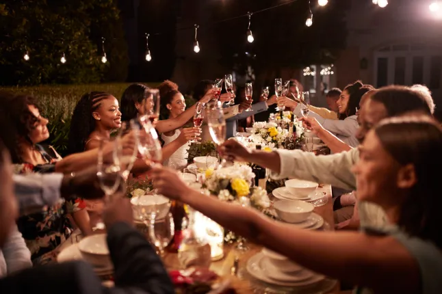 Heureux invités de mariage grillant une boisson tout en étant assis à une table à manger éclairée pendant le dîner de réception